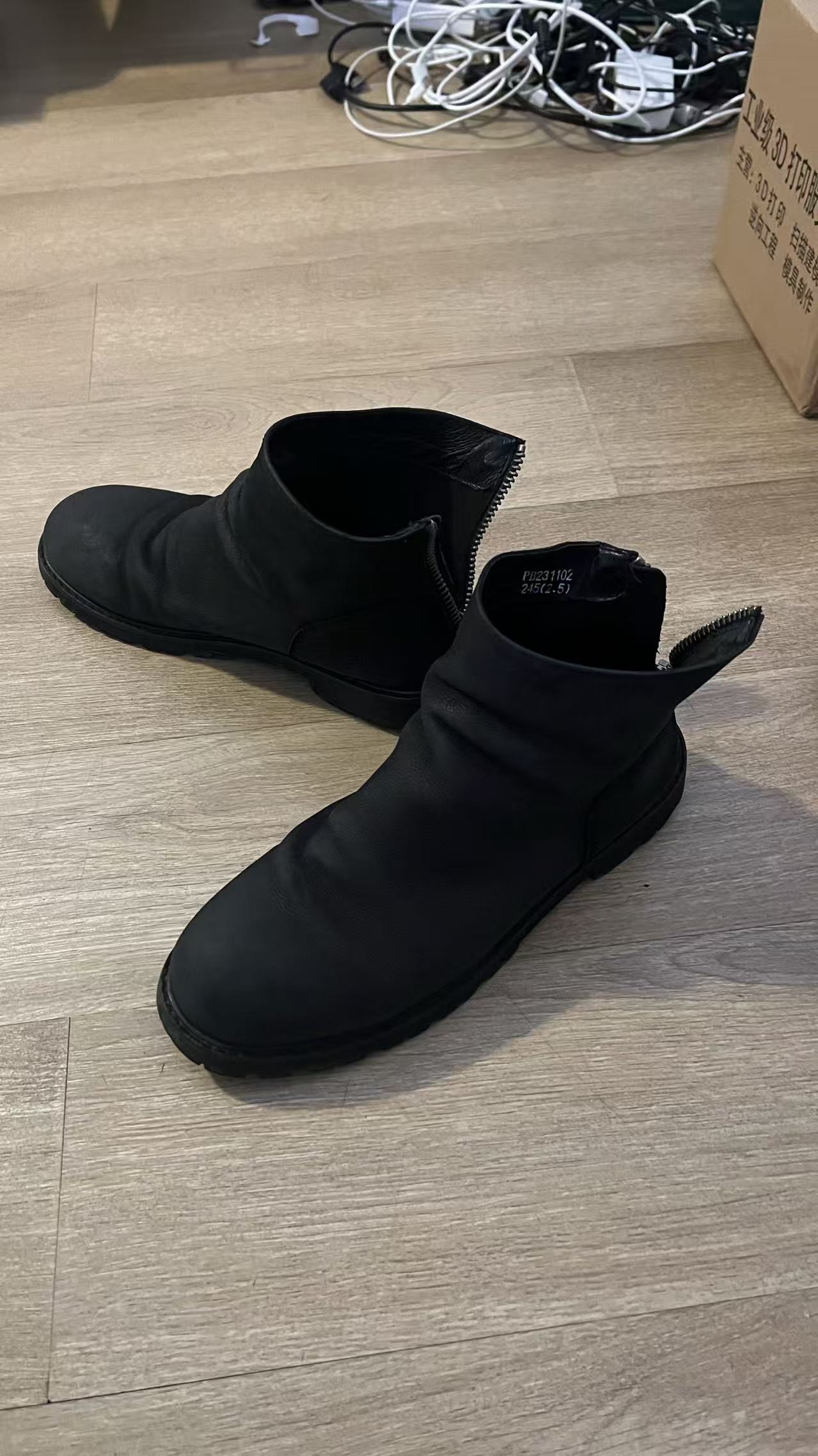 A pair of black leather boots on a wooden floor, shot on a phone, boxes and cables behind them